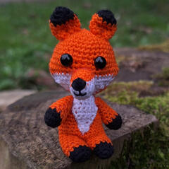 A small crochet fox is sitting on a tree stump and looking into the camera.